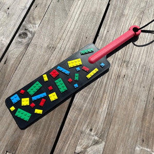 Een BDSM paddle in zwart met gekleurde bouwsteentjes en een rood handvat