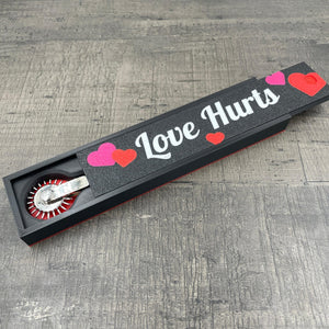 Een silverkleurige BDSM Wartenberg pinwheel in een doosje met de tekst "Love Hurts"