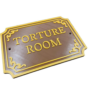 Een bruin deurbordje met de tekst "Torture Room" in gouden letters.